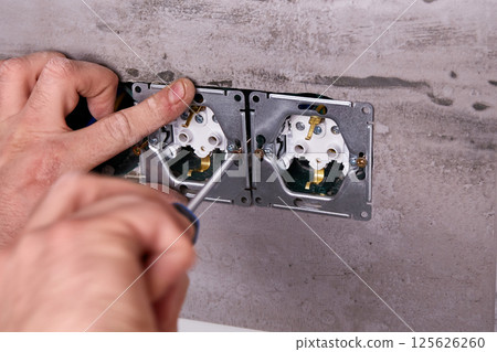 An electrician uses a screwdriver to install an electrical outlet on a wall covered with porcelain stoneware An electrician uses a screwdriver to install an electrical outlet on a wall covered with porcelain stoneware 125626260