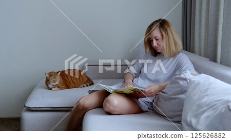 Middle aged woman peacefully reading a book on the sofa at home, with her ginger cat sleeping beside her Middle aged woman peacefully reading a book on the sofa at home, with her ginger cat sleeping beside her 125626882