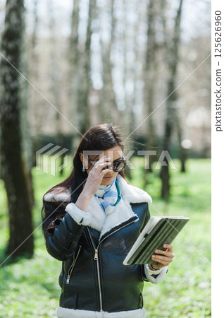 A well-dressed young woman is walking in a park. She is looking seriously at her tablet and touching her glasses.   125626960
