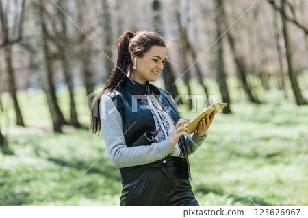 A beautiful girl smiles sincerely and looks at the tablet. Modern young girl wearing headphones with a tablet in a spring park. 125626967