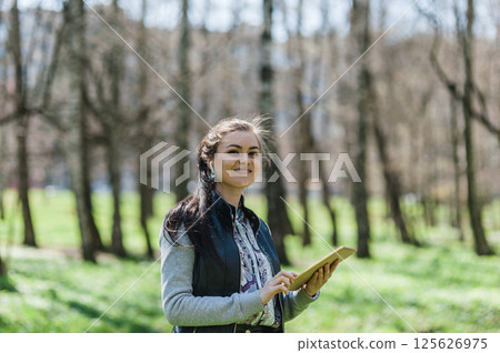 A beautiful girl in leather pants and bomber jacket with a tablet in a spring park. Happy woman with a tablet in her hands smiling sweetly. A beautiful girl in leather pants and bomber jacket with a tablet in a spring park. Happy woman with a tablet in her hands smiling sweetly. 125626975