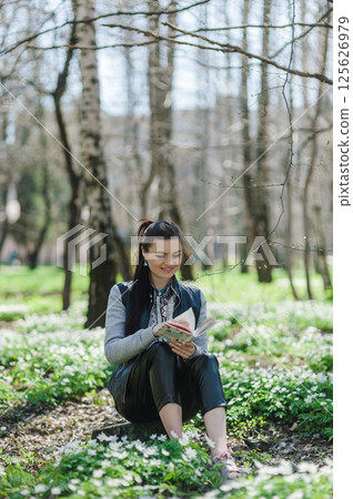 A girl in a spring park resting with a book in her hands. A young woman is sitting on the grass in a green forest.  125626979