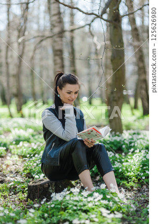 The woman is looking at the book and smiling. A beautiful girl is sitting on a lawn in a park. 125626980