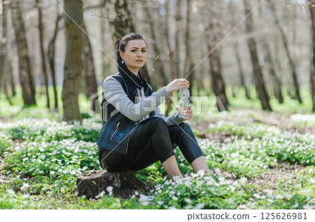 Black-haired woman opens a plastic bottle of water in a spring forest sitting on a tree stump. Beautiful girl in the park with a bottle of water sitting on a tree stump.  125626981
