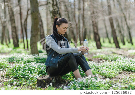 Black-haired woman opens a plastic bottle of water in a spring forest sitting on a tree stump. Beautiful girl in the park with a bottle of water sitting on a tree stump.  125626982