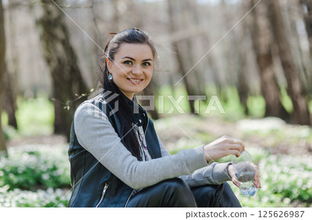 A beautiful girl in leather pants and bomber jacket with a tablet in a spring park. Happy woman with a tablet in her hands smiling sweetly. 125626987