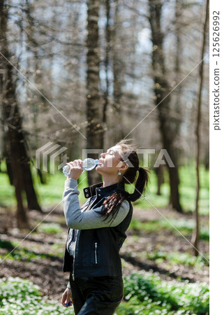 Black-haired woman drinking water from a plastic bottle in the forest. A beautiful girl in a green park drinking water from a bottle. 125626992