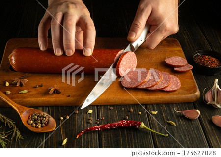 Hands expertly slice into a smoked sausage on a rustic wooden board. Surrounding ingredients include colorful spices, herbs, and garlic, enhancing the culinary experience 125627028