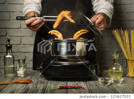 A chef skillfully prepares chicken wings in a pot while steam rises from the cooking process. Various ingredients are visible on the countertop, creating a lively kitchen atmosphere 125627031