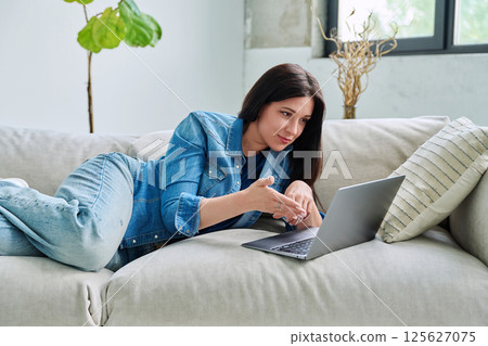 Young woman looking at laptop screen talking having video call lying on couch at home 125627075