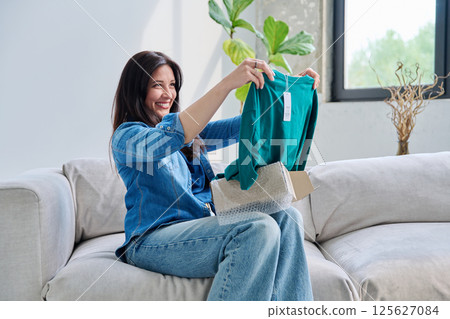 Young woman unpacking cardboard box parcel with online order, looking at new clothes 125627084