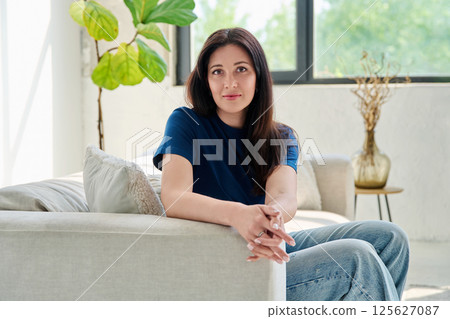 Portrait of beautiful young brunette woman sitting on sofa in living room 125627087