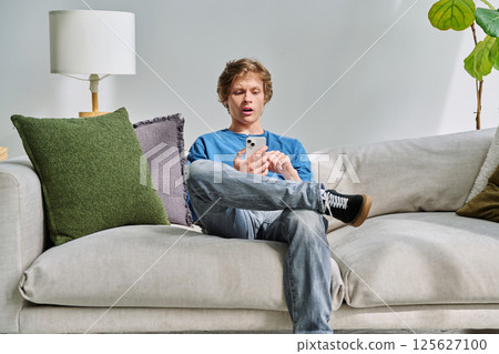 Handsome surprised teenage guy using smartphone sitting on couch at home Handsome surprised teenage guy using smartphone sitting on couch at home 125627100