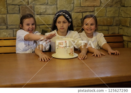 Smiling girls and a vibrant cake create a perfect festive moment. The sisters gently lean toward the dessert, admiring each detail with delight. 125627290
