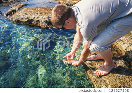 A young man releases a fish into the water on a rocky riverbank 125627398