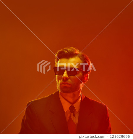 Man in sunglasses with confident chin-up pose against deep orange glow expressing cold detachment 125629696