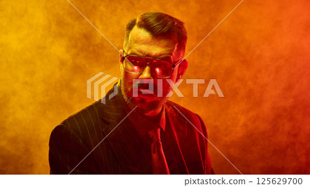 Bearded man in orange-red neon light and fog looks sideways with dramatic tension across face reflecting emotional conflict. 125629700