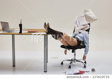 Young woman buried in papers with legs on desk during surreal office moment against white studio background. 125629710