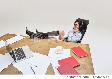 Female employee relaxing in chair with legs on desk while talking on headphones and smiling against white background. 125629713