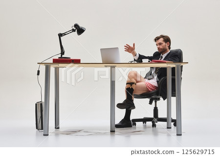 Young handsome, confident man gesturing during virtual meeting while dressed in business suit and underwear from waist up. Young handsome, confident man gesturing during virtual meeting while dressed in business suit and underwear from waist up. 125629715