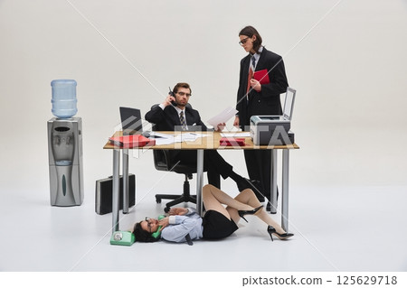 Two men in suits stand near desk while female worker talks on phone lying under table in strange office scene. Horizontal photo. Two men in suits stand near desk while female worker talks on phone lying under table in strange office scene. Horizontal photo. 125629718