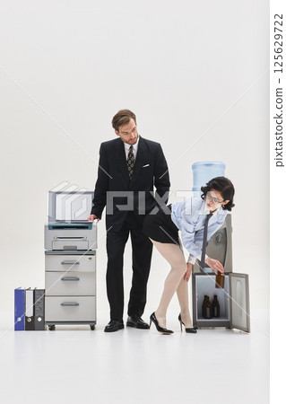 Man in suit looking at woman bending near water cooler while taking bottle from fridge in surreal office setup. Man in suit looking at woman bending near water cooler while taking bottle from fridge in surreal office setup. 125629722