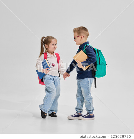 Schoolboy and schoolgirl holding hands with backpacks and books in school outfits walk together against studio background. 125629750