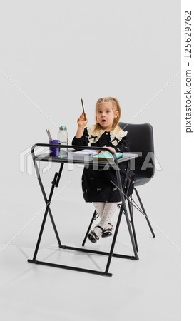 Young girl in black dress sitting at desk raising hand with curious facial expression and open notebooks. Concept of active learning 125629762