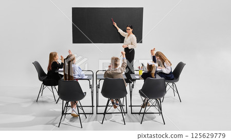 Teacher pointing at blackboard as all children actively raise hands to answer question. Concept of interactive learning Teacher pointing at blackboard as all children actively raise hands to answer question. Concept of interactive learning 125629779