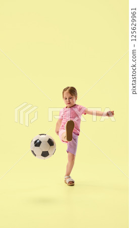 Focused girl in pink raising leg and kicking soccer ball with precision and balance against yellow studio background. Focused girl in pink raising leg and kicking soccer ball with precision and balance against yellow studio background. 125629961