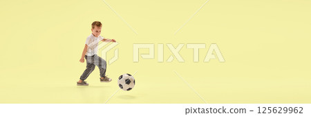 Banner. Athletic young boy kicks soccer ball forward in motion while running across yellow studio background with negative space. 125629962
