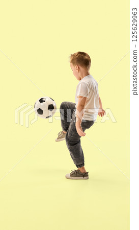 Little champion, sporty boy balances soccer ball with foot focused on action against yellow studio background showing coordination. 125629963