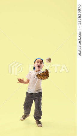 Excited boy, focused on game, dressed in cap catches baseball with glove smiling against yellow studio background. 125629970
