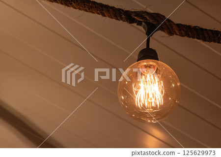 Warm glowing Edison-style vintage filament bulb hanging from rustic rope fixture 125629973
