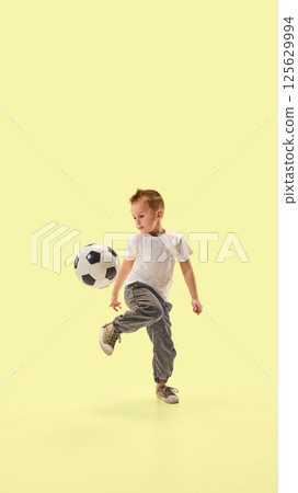 Young boy skillfully kicks soccer ball with raised leg showing control and motion in active pose. Child training against yellow background. 125629994