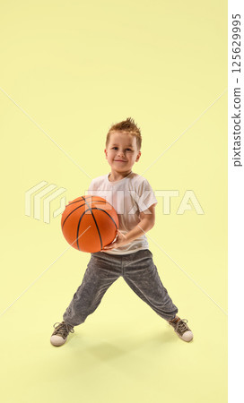 Smiling boy dressed sportwear, posing with basketball in wide stance against yellow background showing joy and playfulness. Smiling boy dressed sportwear, posing with basketball in wide stance against yellow background showing joy and playfulness. 125629995