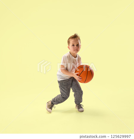 Focused little boy, junior champion with basketball prepares to dribble showing determination under against yellow studio background. 125629997