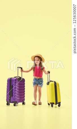 Full length photo of little charming girl, dressed stylish summer attire and hat, pulling suitcases against pastel yellow background. 125630007