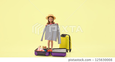Little girl in summer hat packing suitcase with clothes and toys against pastel studio background preparing for travel adventure. 125630008