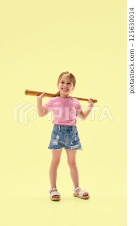 Smiling little girl holding wooden bat behind shoulders standing relaxed and showing confidence against yellow background. Smiling little girl holding wooden bat behind shoulders standing relaxed and showing confidence against yellow background. 125630014