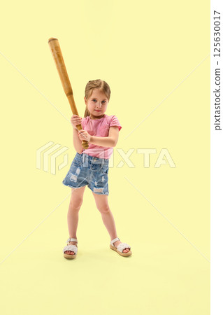 Focused preschooler, stylish girl holding wooden bat in standing pose with confident look against pastel yellow background. Focused preschooler, stylish girl holding wooden bat in standing pose with confident look against pastel yellow background. 125630017