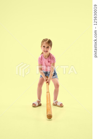 Full length photo of happy girl with pink shirt swinging wooden bat showing joy and energy against pastel yellow background. 125630019