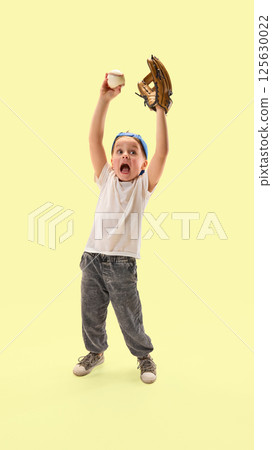 Happy boy proudly holds baseball glove and ball high over head in victory gesture against yellow studio background. Happy boy proudly holds baseball glove and ball high over head in victory gesture against yellow studio background. 125630022