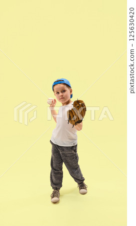 Little boy holding glove and baseball preparing to throw against pastel yellow background. Junior champion training. Little boy holding glove and baseball preparing to throw against pastel yellow background. Junior champion training. 125630420