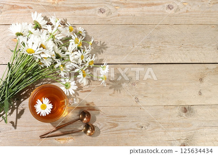 Chamomile tea in a transparent cup with natural small chamomile flowers on a light rustic table, the concept of herbal tea and proper natural nutrition, summer postcard, selective focus 125634434