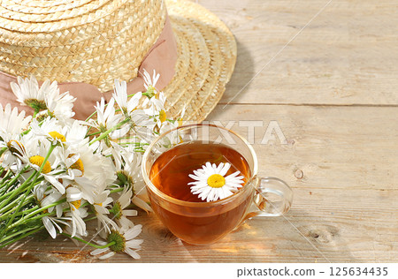 Chamomile tea in a transparent cup with natural small chamomile flowers and a summer hat on a light rustic table, the concept of herbal tea and proper natural nutrition, selective focus Chamomile tea in a transparent cup with natural small chamomile flowers and a summer hat on a light rustic table, the concept of herbal tea and proper natural nutrition, selective focus 125634435