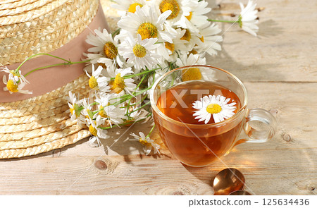 Chamomile tea in a transparent cup with natural small chamomile flowers and a summer hat on a light rustic table, the concept of herbal tea and proper natural nutrition, selective focus 125634436