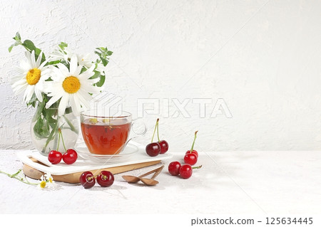 Chamomile morning tea in a transparent cup with natural small chamomile flowers, mint and fresh cherry berries on a light sunny table, the concept of herbal tea and proper natural nutrition, promote 125634445