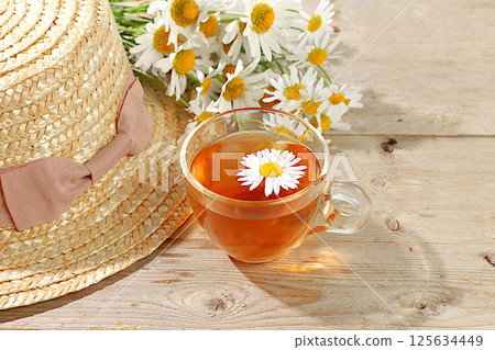 Chamomile tea in a transparent cup with natural small chamomile flowers on a light rustic table, concept of herbal tea and proper natural nutrition, summer card and outdoor recreation, selective focus 125634449