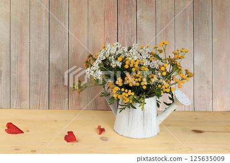 Tansy and yarrow in a watering can on a wooden background, collection of useful herbs for treatment according to folk recipes. Floral arrangement of wild herbs and flowers on a wooden table, rustic 125635009